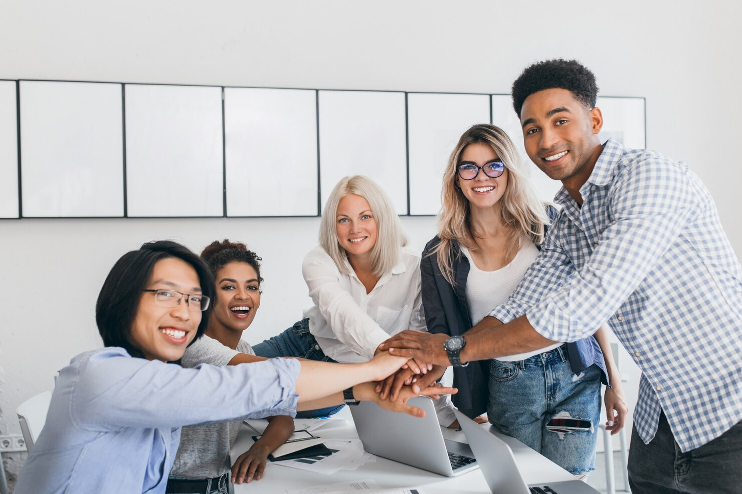 A group of ethnically diverse people gather around a desk of computers and overlap hands in a moment of team comradery.